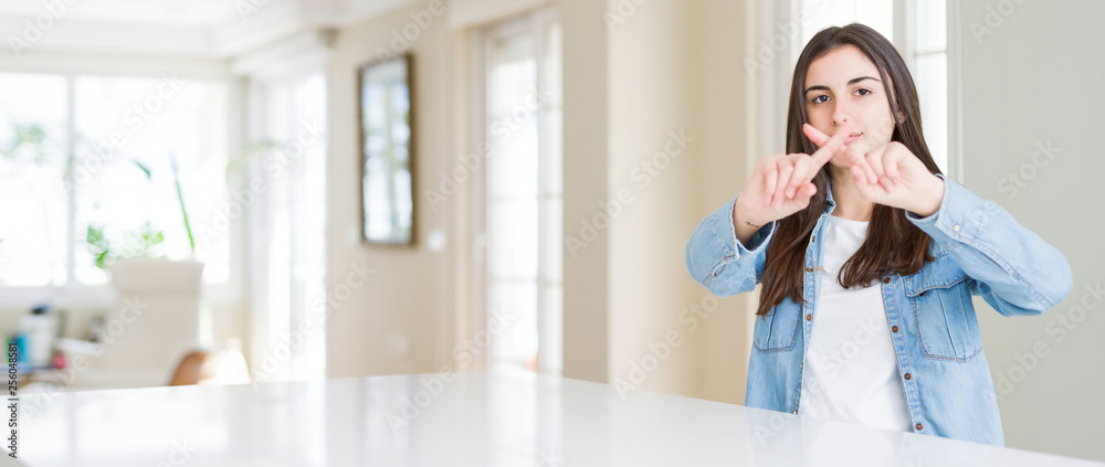 Wide angle picture of beautiful young woman sitting on white table at home Rejection expression crossing fingers doing negative sign Wide angle picture of beautiful young woman sitting on white table at home Rejection expression crossing fingers doing negative sign