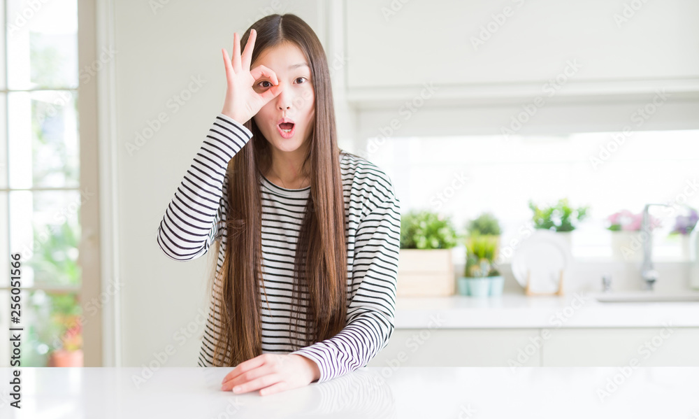 Beautiful Asian woman wearing stripes sweater doing ok gesture shocked with surprised face, eye looking through fingers. Unbelieving expression.