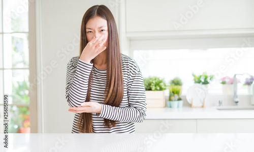 Beautiful Asian woman wearing stripes sweater smelling something stinky and disgusting  intolerable smell  holding breath with fingers on nose. Bad smells concept.