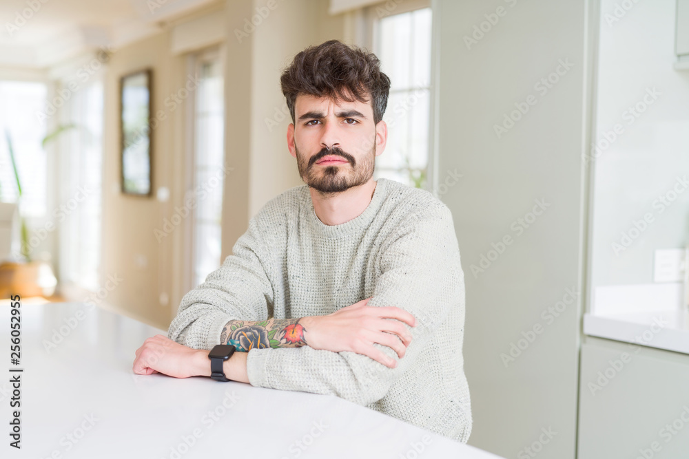 Young man wearing casual sweater sitting on white table skeptic and nervous, disapproving expression on face with crossed arms. Negative person.