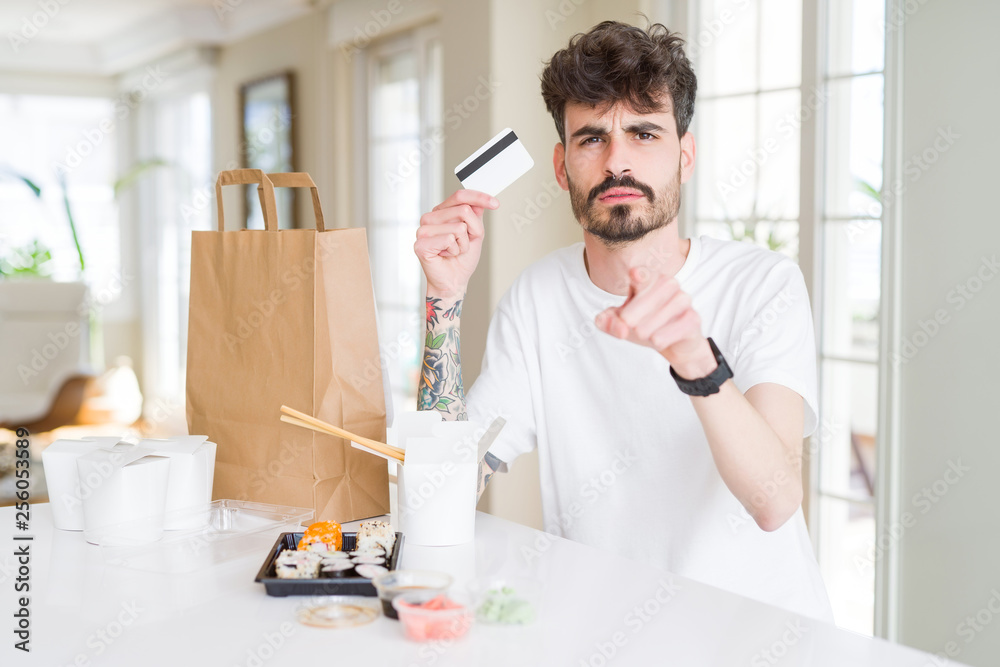 Young man eating asian sushi from home delivery using credit card as payment pointing with finger to the camera and to you, hand sign, positive and confident gesture from the front