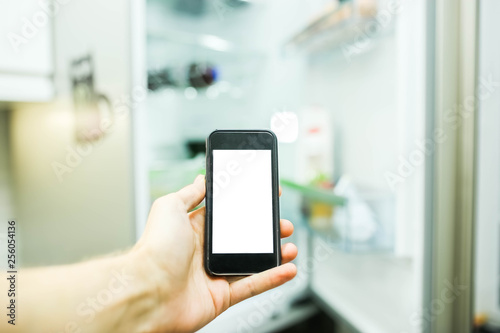 Smartphone with a white screen on the blurred background of an open refrigerator with products in the kitchen. Smart home technology system.