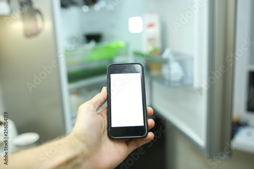 Smartphone with a white screen on the blurred background of an open refrigerator with products in the kitchen. Smart home technology system.