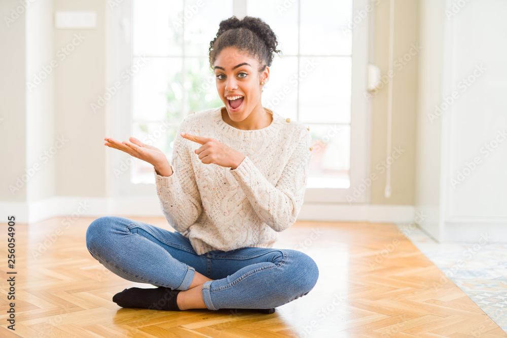Beautiful young african american woman with afro hair sitting on the floor amazed and smiling to the camera while presenting with hand and pointing with finger. Beautiful young african american woman with afro hair sitting on the floor amazed and smiling to the camera while presenting with hand and pointing with finger.