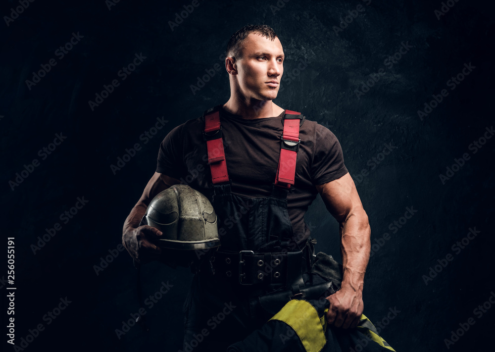 Portrait of a brutal muscular fireman holding a helmet and jacket standing in the studio against a dark textured wall