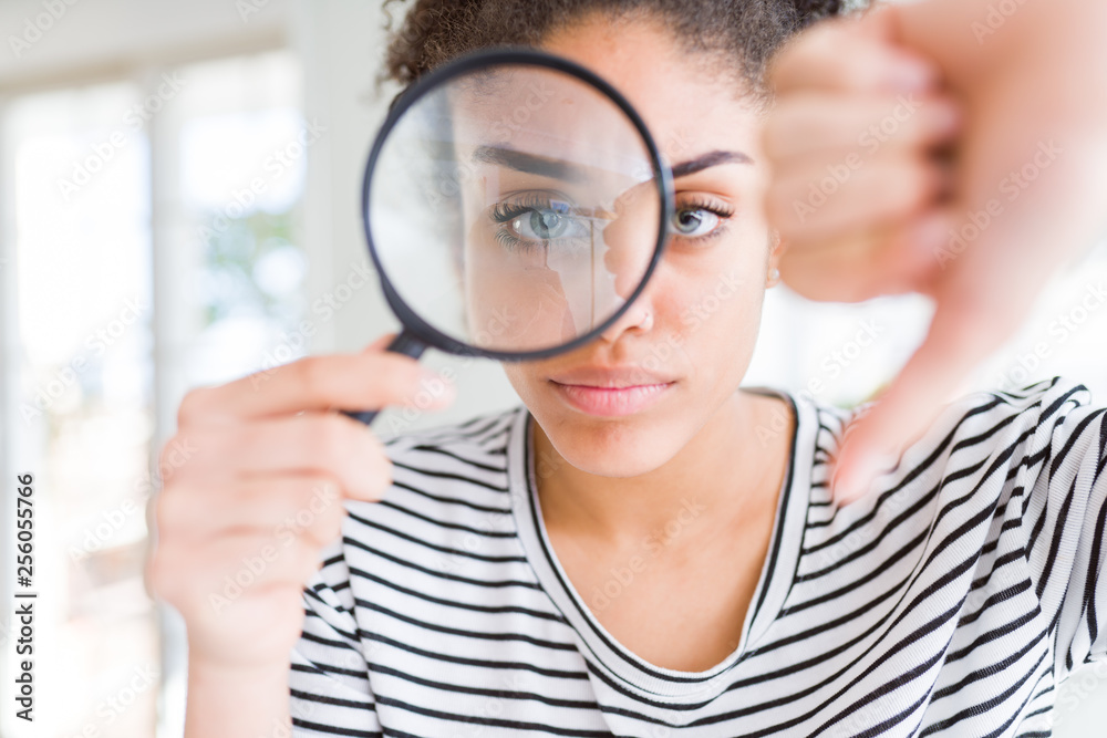 Young african american woman looking through magnifying glass with angry face, negative sign showing dislike with thumbs down, rejection concept