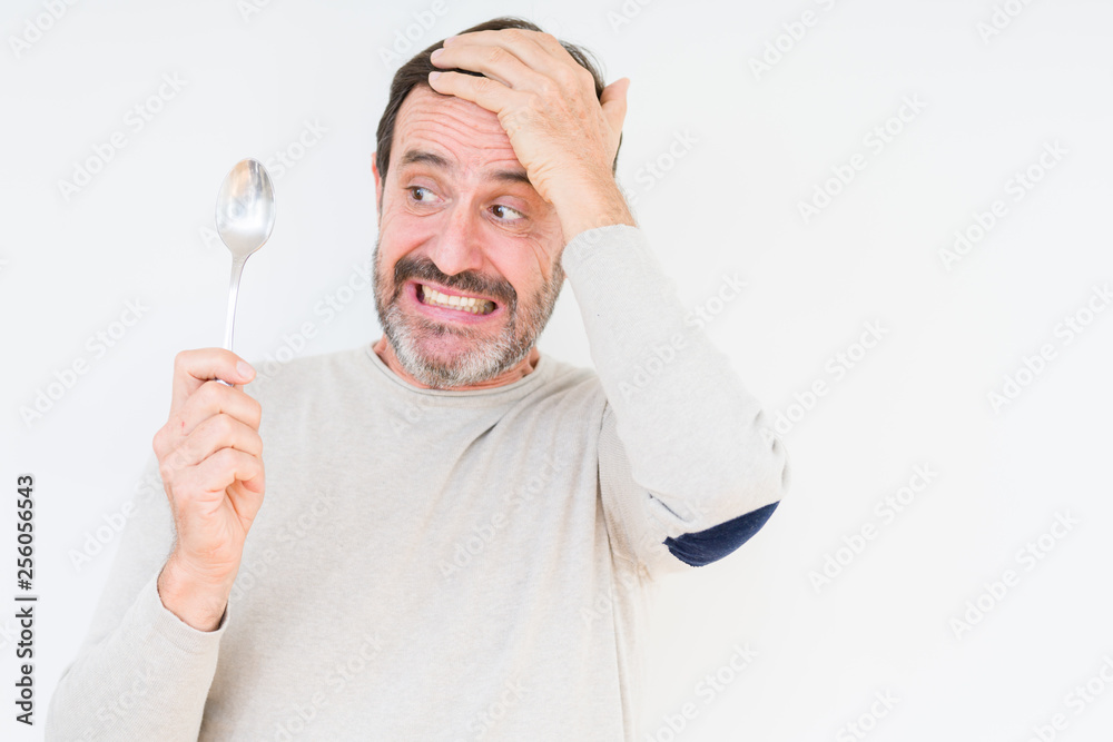 Senior man holding silver spoon over isolated background stressed with hand on head, shocked with shame and surprise face, angry and frustrated. Fear and upset for mistake. Senior man holding silver spoon over isolated background stressed with hand on head, shocked with shame and surprise face, angry and frustrated. Fear and upset for mistake.