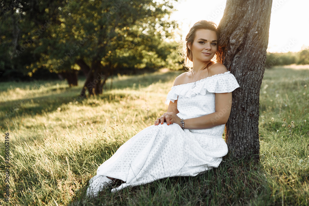 Gorgeous smiling girl sitting on the grass near the tree during sunset in white modern sundress Gorgeous smiling girl sitting on the grass near the tree during sunset in white modern sundress