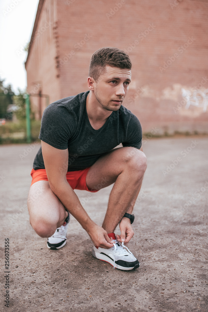 Athlete man crouched, tying shoelaces on sneakers. In the summer on the sports ground in the city. Trainer guy in the watch tracker bracelet after jogging. Workout outdoor lifestyle.