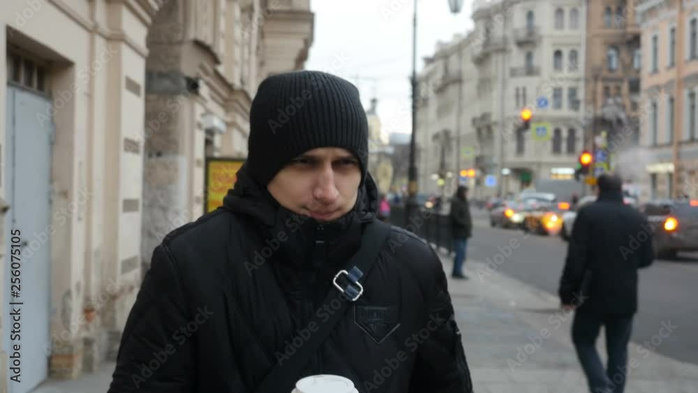 Front view of young man walking down the city street and drinking coffee. Serious tired man in black walks on the street. Front view of young man walking down the city street and drinking coffee. Serious tired man in black walks on the street.