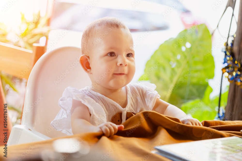 Fototapeta premium Infant girl is sitting on a baby high chair in a street cafe by the table. Children in white dress dabbles and pulls the tablecloth