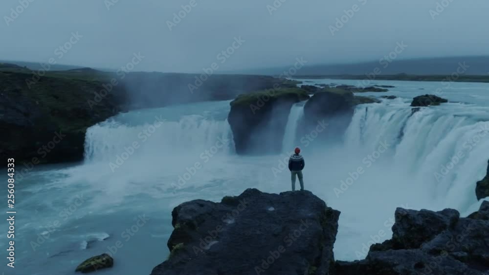Epic adventure seeker drone shot of urban nomad traveller,hipster young millennial man stand on edge of cliff near waterfall in iceland, confident and brave, search for new exciting horizons and ideas