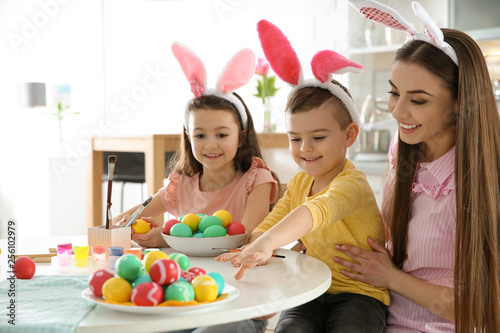 Mother and her children with bunny ears headbands painting Easter eggs in kitchen, space for text