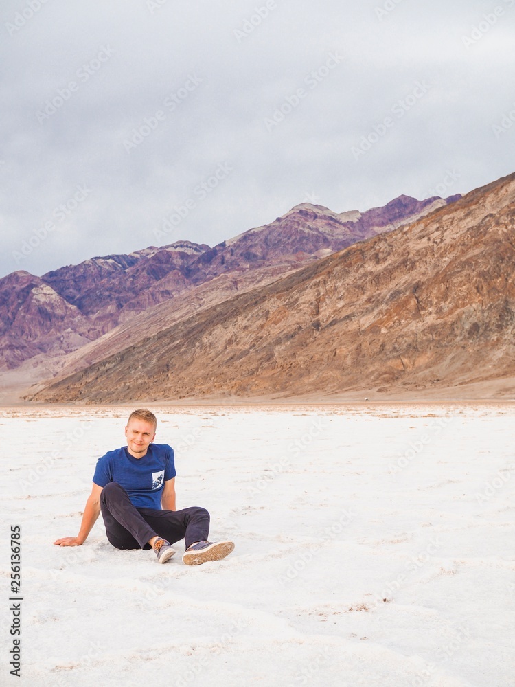 The young blond man in a blue shirt and shorts is sitting on a saline soil Badwater in Death Valley