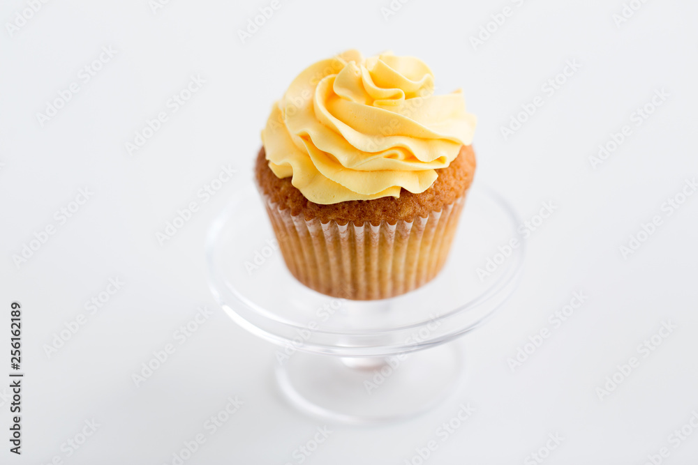 food, pastry and sweets concept - cupcake with yellow buttercream frosting on glass confectionery stand over white background food, pastry and sweets concept - cupcake with yellow buttercream frosting on glass confectionery stand over white background
