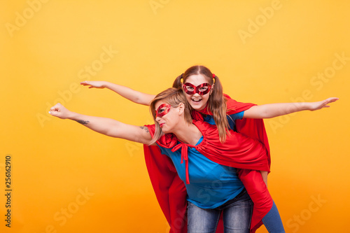 Beautiful mother and her cute little girl playing together dressed like superheros over yellow background.
