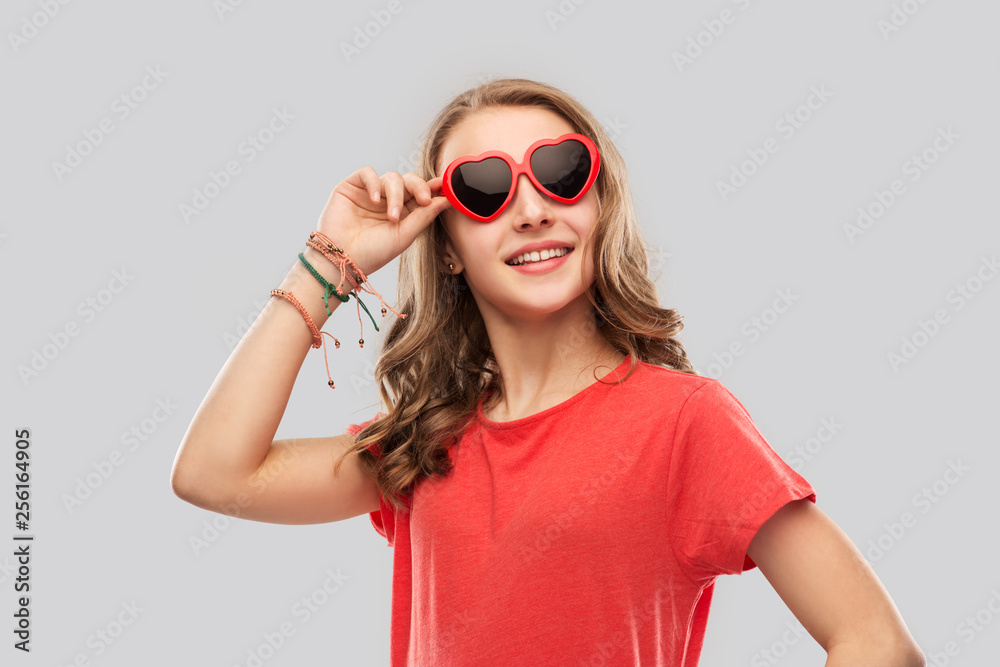 Fototapeta premium valentine's day, love and people concept - smiling pretty teenage girl wearing bangles on hand in red heart shaped sunglasses over grey background