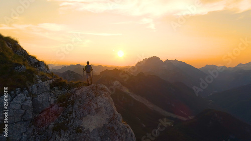 DRONE: Flying around a male hiker standing on the mountain peak at sunset.