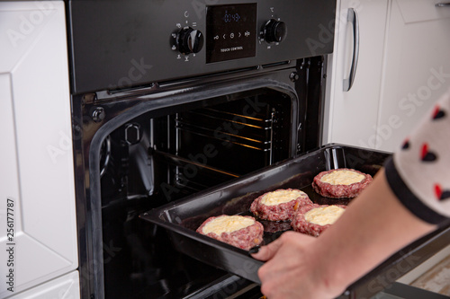 Woman hands putting baking tray with cutlets or meatballs and into oven