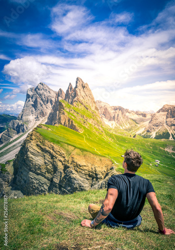 vertical shot of a beautiful young muscular man sitting on the grass enjoying the beautiful dolomites mountains landscape. seceda, 2500m over the odle group mountains