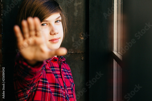 Portrait thoughtful caucasian teenager with long straight hairstyle showing stop sign with palm, looking at camera in dark modern loft interior. Child wthout parents, independent concept.