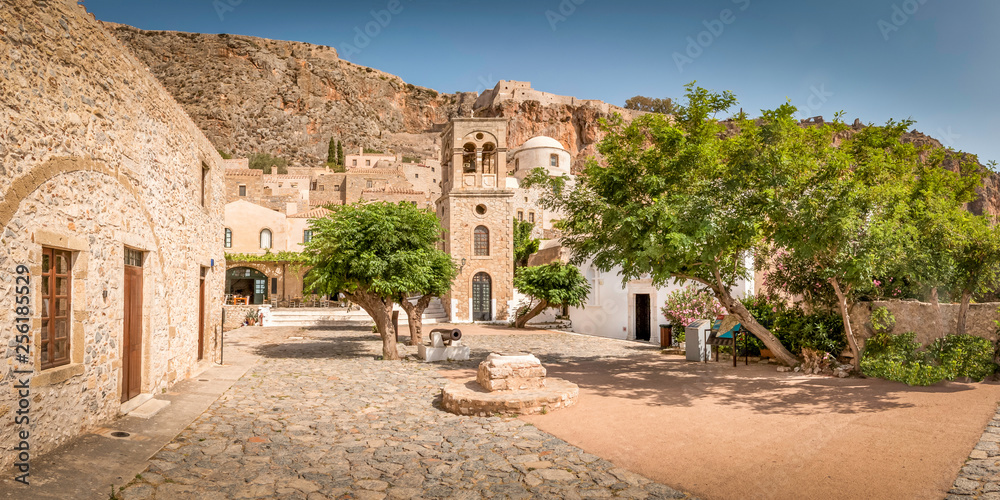 Sunny village square with some trees and an old fortress on top of the hills in the middle of Monemvasia, Peleponnese, Greece