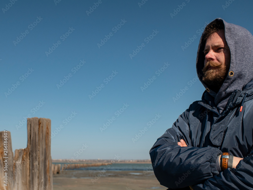 man hipster with a beard and mustache in the hood against the blue sky and estuary
