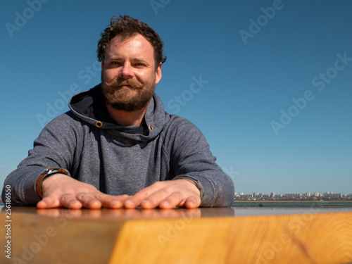 funny man with a beard and mustache sitting at a coffee table against a blue sky © yavorovich