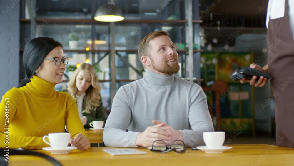 Cheerful caucasian man and young Asian woman smiling and talking with waiter while paying with smartphone on lunch in the restaurant Cheerful caucasian man and young Asian woman smiling and talking with waiter while paying with smartphone on lunch in the restaurant