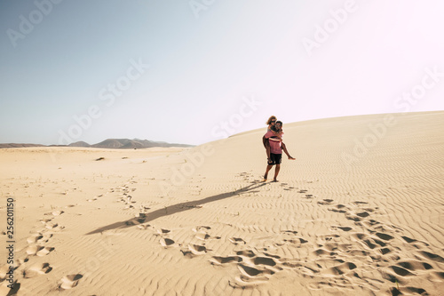 Happy traveler couple enjoy the desert dunes carrying eachother with love and fun - Caucasian people tourists walk together on the beach in outdoor leisure activity - clear sky