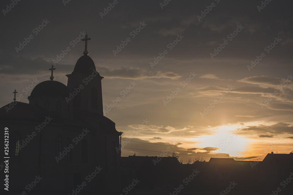 Orthodox church silhouette at sunset with cloudy sky and sun in background.
