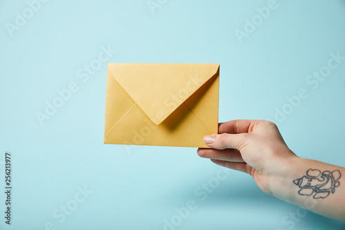 partial view of woman with tattoo holding yellow envelope on blue background