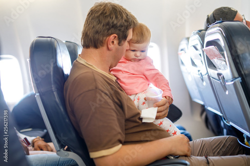 Young tired father and his crying baby daughter during flight on airplane going on vacations. Dad holding and playing with baby girl on arm. Air travel with baby, child and family concept