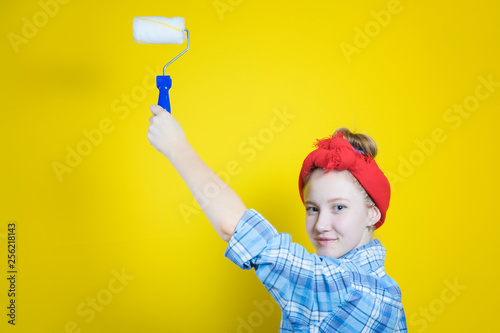 Young beautiful girl in a plaid shirt and a red bandage on his head, paints with a paint roller against the background of a yellow studio. Retro style. photo