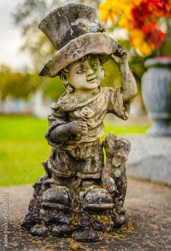 A aged alabaster boy grave statue on a grave. The boy is holding his hat, and there is also a dog t as well by his leg. The alabaster has discolored from the elements. 