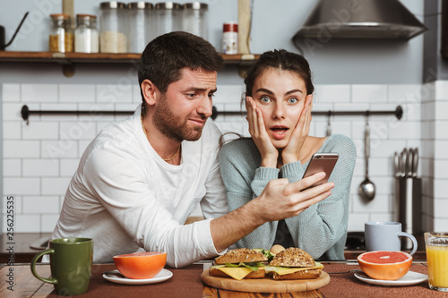 Shocked young loving couple at the kitchen have a breakfast using mobile phone.