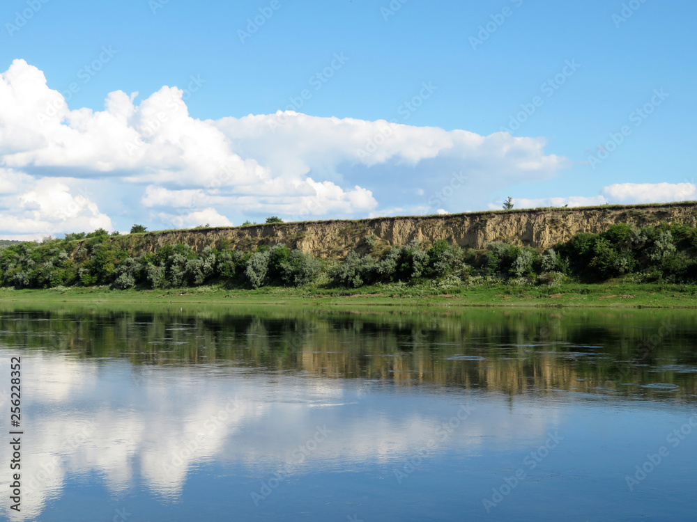 Picturesque landscape with green river bank, rocky hills and blue sky. Rural scene in summer, white clouds are reflected in the water surface