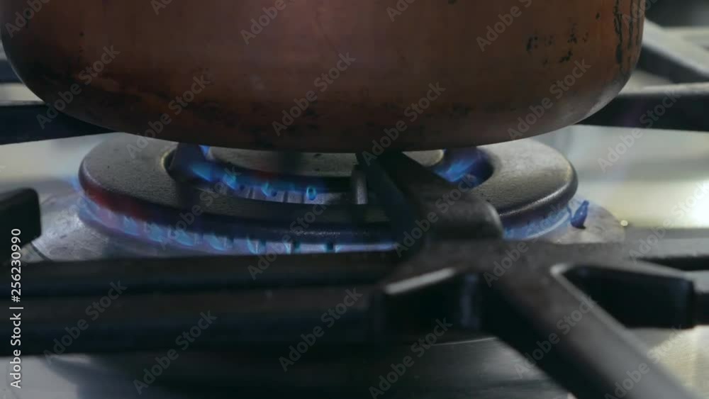 Zoom out of close shot of a burner getting lit with blue flames on a gas stove and a golden brass pot on it. Zoom out of close shot of a burner getting lit with blue flames on a gas stove and a golden brass pot on it.