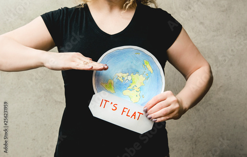  Flat Earther concept. Person who believes that Earth is flat disc. Anonymous woman holding flat Earth model in front of body with text: It`s flat. Isolated on gray background, studio shot.  photo