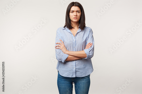 Indoor photo of brunette woman, looks with distrust wary suspicion, tense, listens to someone with doubt, stressful, standing with arms folded dressed in jeans and striped shirt, over white wall