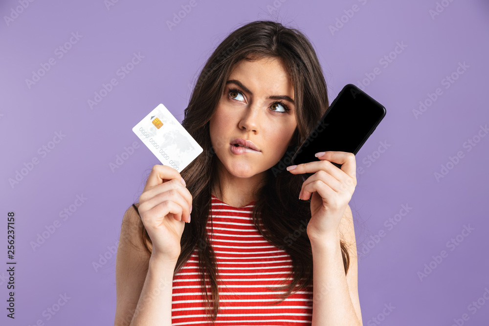 Serious young pretty woman posing isolated over purple background wall using mobile phone holding credit card.