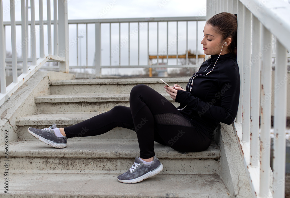 Young woman in black sports outfit resting and listening music on smartphone after running in the city.