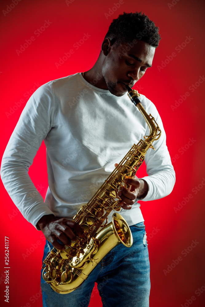 Naklejka premium African American handsome jazz musician playing the saxophone in the studio on a neon background. Music concept. Young joyful attractive guy improvising. Close-up retro portrait.
