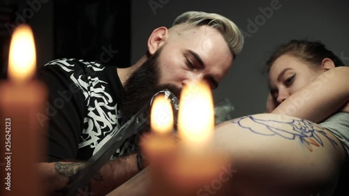closeup candles turn on foreground and tattoo master with thick black beard creates tattoo on young girl thigh