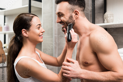 Schilderij op canvas attractive woman shaving face of handsome shirtless boyfriend in bathroom