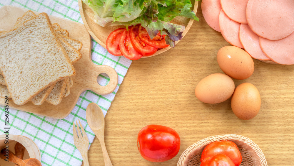 Homemade sandwich breakfast preparing. Whole wheat bread on wooden cutting board on white fabric. Slice tomatoes and lettuce on wooden plate. Fresh tomatoes, ham, eggs, spoon and fork on wooden table.