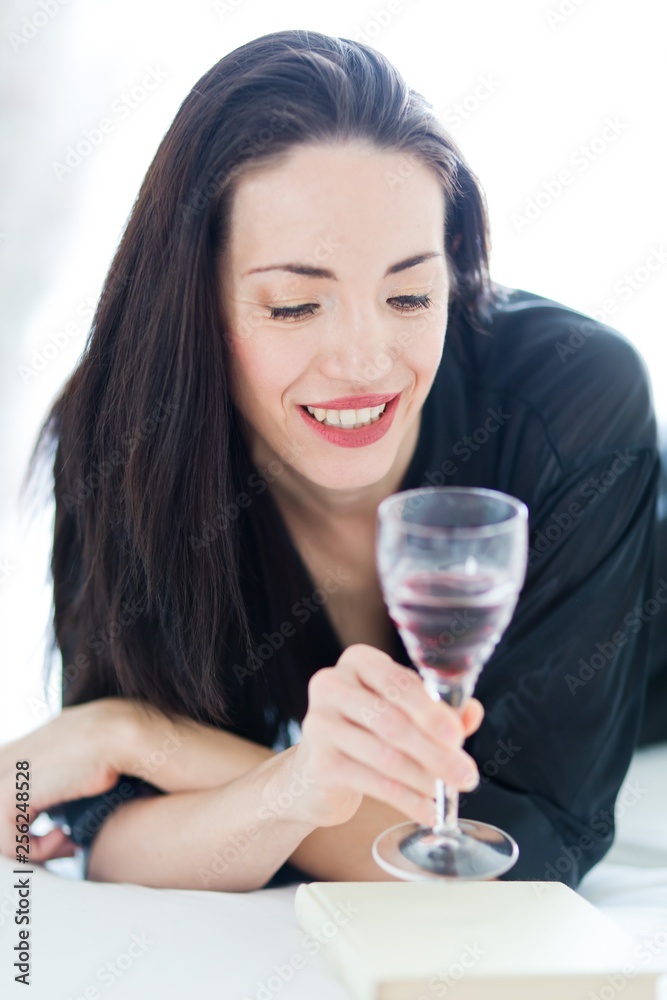 Pretty woman in black laying on bed and looking into glass of wine.