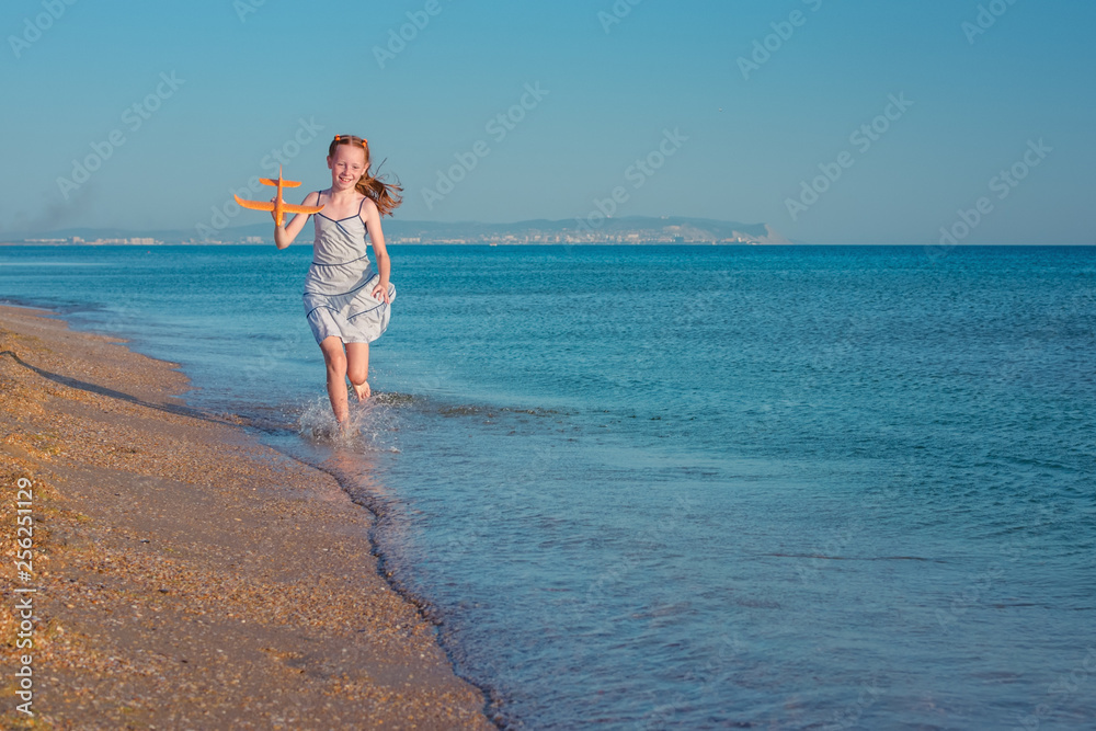 Cheerful red-haired girl playing with toy airplane and jumps over the waves of the sandy shore of the sea against the background blue summer sky. Rejoices in the rays of the sun and freedom. Cheerful red-haired girl playing with toy airplane and jumps over the waves of the sandy shore of the sea against the background blue summer sky. Rejoices in the rays of the sun and freedom.
