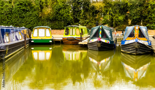 Public view of five colourful, unoccupied canal boats at their moorings in Sovereign Wharf, Banbury. Reflections of the vessels in water. Hedgerow background. Oxford canal waterway. England.