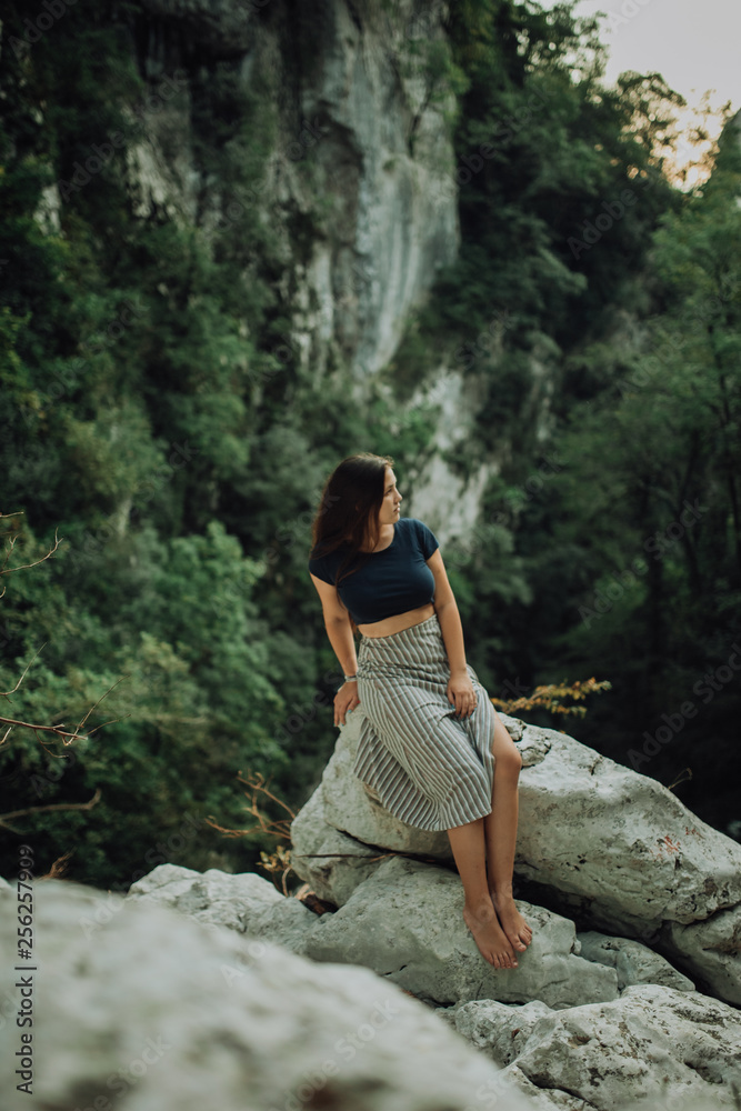 Young and sexy woman sit on rock wearing swimsuit on the beautiful waterfall in the jungle beside cliff